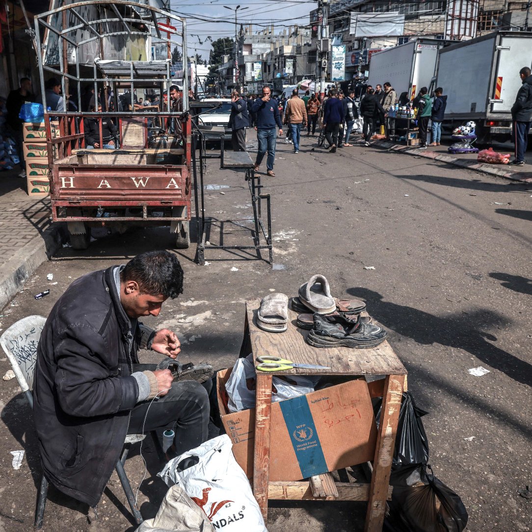 در تصویر، مردی در حال تعمیر یا واکس زدن کفش‌ها است. او روی یک صندلی نشسته و وسایل کارش را بر روی یک میز کوچک چوبی قرار داده است. در اطراف او، افرادی در حال عبور و مرور هستند و تعدادی خودروی باربری در پس‌زمینه دیده می‌شود. فضایی شلوغ و شهری به نظر می‌رسد و فعالیت‌های روزمره در حال انجام است. چند کشیده و بقیه اجناس نیز در اطراف قرار دارند.