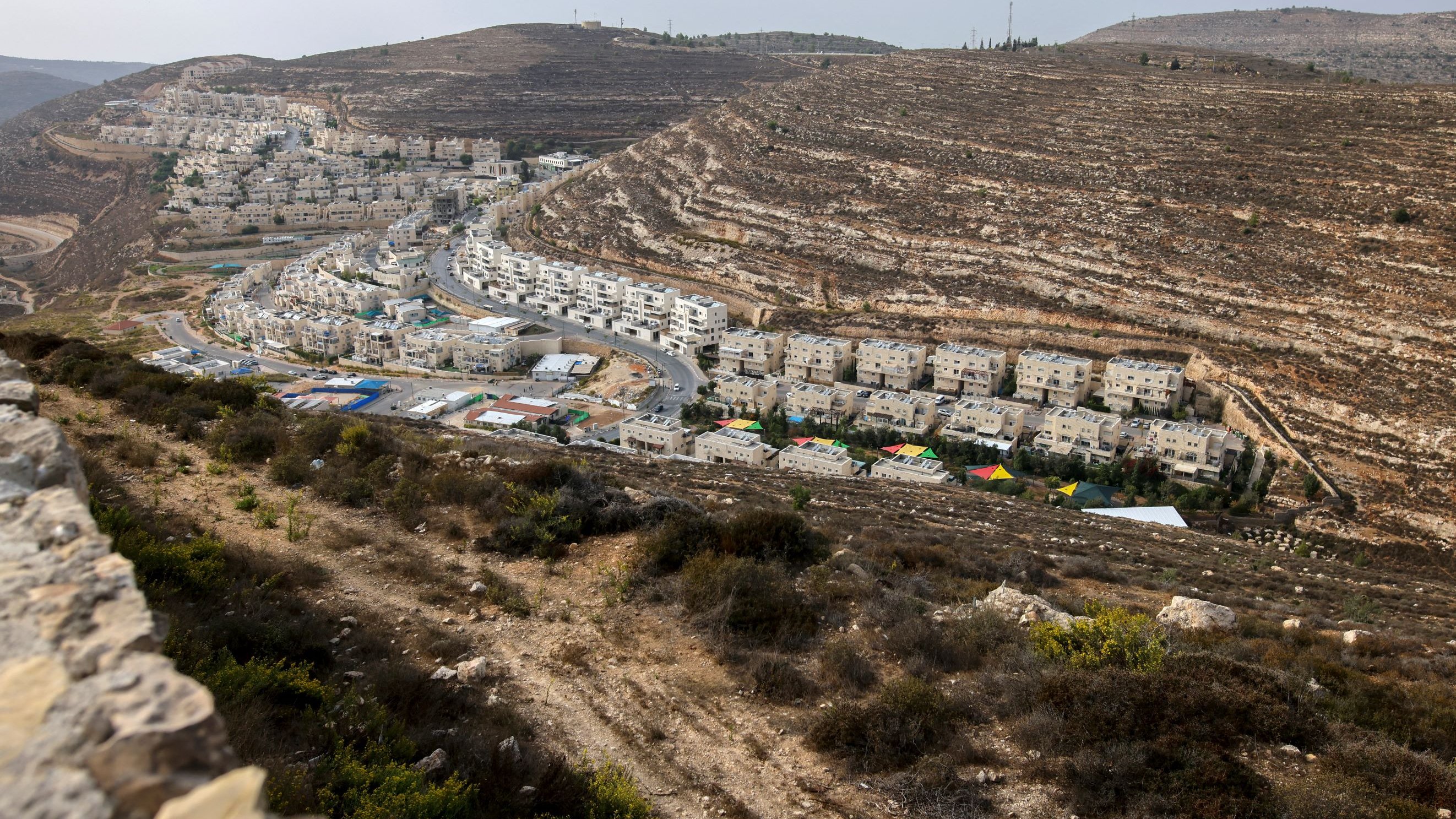 L'image montre un paysage vallonné avec des bâtiments résidentiels. On voit des maisons qui sont construites en terrasses sur les pentes de la colline, ainsi qu'une route qui serpente à travers l'urbanisation. La végétation est sparse et composée de buissons et d'herbes. Au fond, on aperçoit d'autres constructions, indiquant une zone développée et habitée. Le ciel est légèrement nuageux, ce qui donne une ambiance un peu grise à l'ensemble.