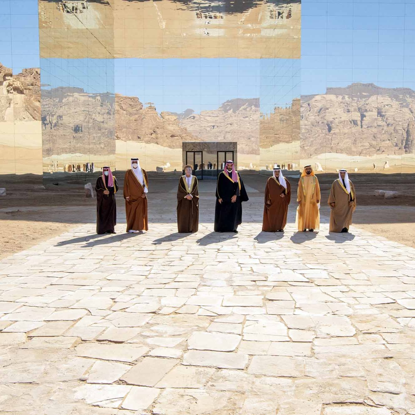 در این تصویر، گروهی از مردان در لباس‌های سنتی عربی در یک فضای باز ایستاده‌اند. پس‌زمینه شامل کوه‌ها و زمین‌های بیابانی است که با آینه‌های بزرگ منعکس می‌شوند. این آینه‌ها نمای جالبی از محیط را ایجاد کرده و بر جذابیت تصویر می‌افزایند. مردان در کنار یکدیگر و به طور منظم ایستاده‌اند و در زمینه خاصی از سنگ‌فرش قرار دارند.