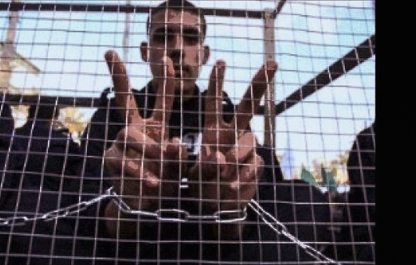 الصورة تظهر شابًا يقف خلف شبكة أو قفص، حيث يرفع يديه مكونًا علامة أو عددًا معينًا بإشارات أصابعه. يبدو أنه محاط بمساحة ضيقة، مما يعطي إيحاءً بالاحتجاز أو العزلة. تعكس تعابير وجهه أو وضع يديه مشاعر معينة، مثل المقاومة أو الإصرار. الخلفية غير واضحة تمامًا، مما يركز الانتباه على الشاب وإشارته.