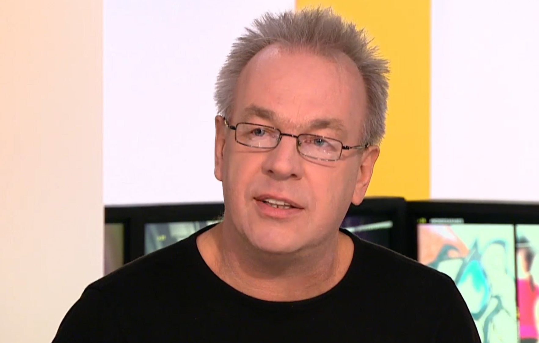 L'image montre un homme avec des cheveux gris et des lunettes. Il porte un t-shirt noir et semble en train de parler, avec un fond flou qui suggère un environnement de discussion ou d'interview.