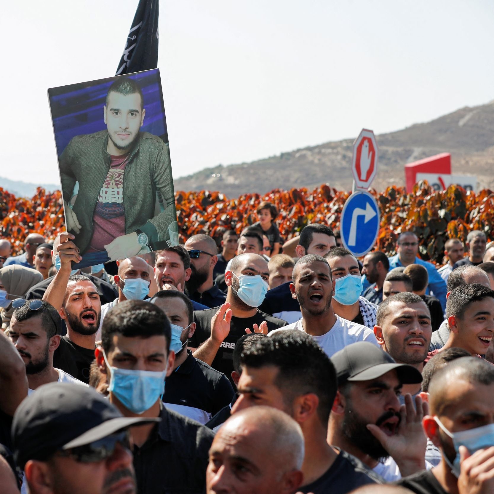 در تصویر، گروهی از افراد در یک تظاهرات حضور دارند. تعداد زیادی از افراد پرچم‌هایی به رنگ‌های مختلف در دست دارند. یکی از تظاهرکنندگان تصویری از یک مرد را بالا نگه داشته است. بیشتر افراد ماسک به صورت دارند و در پس‌زمینه، زمین‌های سرسبز و تپه‌هایی دیده می‌شود. به نظر می‌رسد که این تجمع به منظور اعتراض یا یادبود برگزار شده باشد.