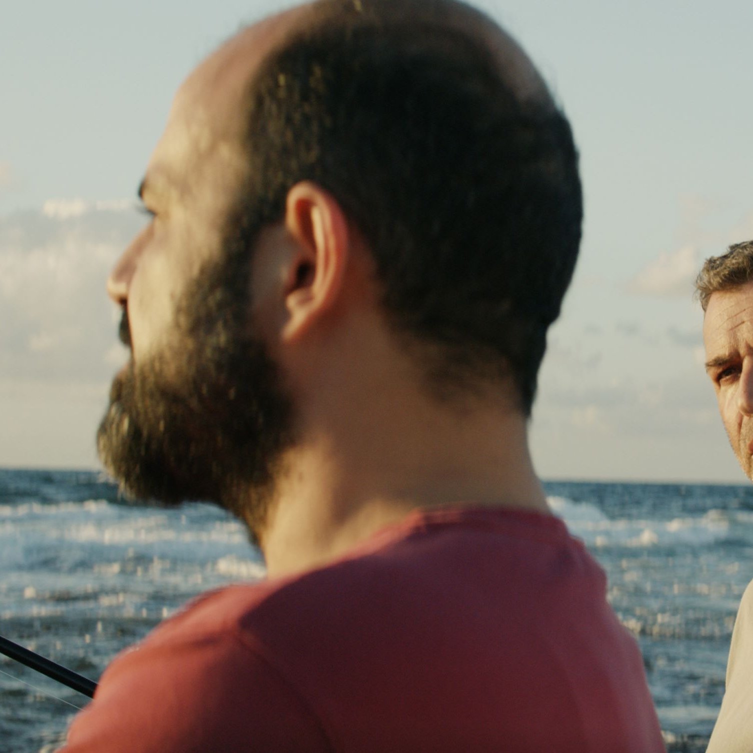 L'image montre deux hommes se tenant près de la mer. L'un d'eux a une barbe et porte un t-shirt rouge, tandis que l'autre a les cheveux grisonnants et un t-shirt clair. L'arrière-plan présente un océan calme sous un ciel bleu parsemé de nuages. L'atmosphère semble sereine, mais les expressions des hommes suggèrent une conversation intense ou réfléchie.