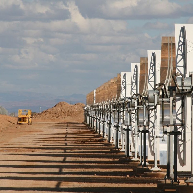 L'image montre une série de panneaux solaires en concentrateurs, alignés sur un terrain désertique. Ces structures lumineuses captent la lumière du soleil pour générer de l'énergie. En arrière-plan, on peut voir un ciel partiellement nuageux avec des montagnes lointaines. L'environnement semble aride, avec des collines de terre et un véhicule de construction visible à distance. Les ombres des panneaux s'étendent sur le sol, soulignant leur orientation vers le soleil.