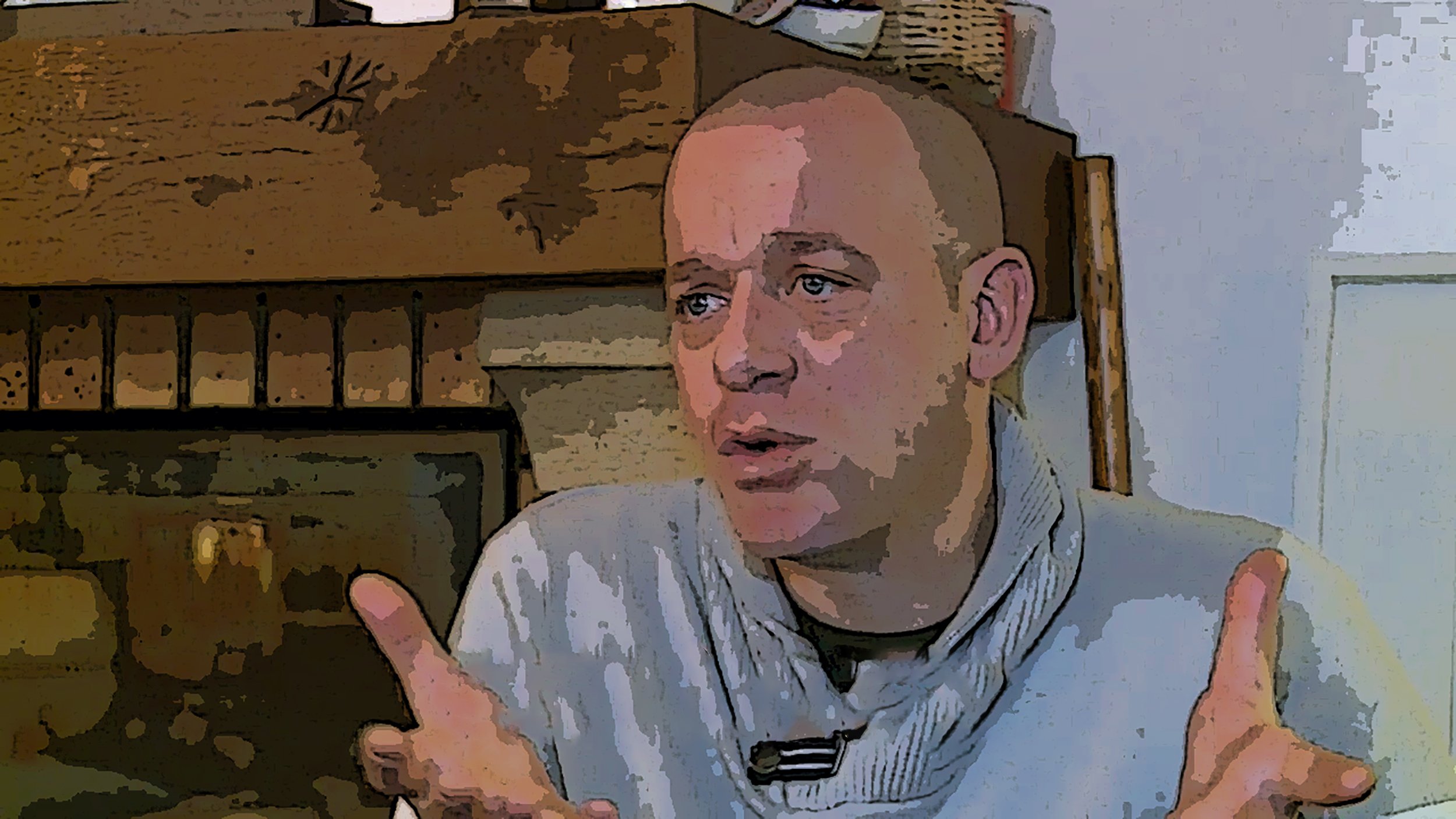 L'image montre un homme avec une coupe de cheveux courte et un pull en tricot. Il est assis près d'une cheminée en pierre. Son expression faciale semble être sérieuse, et il gesticule avec ses mains, ce qui suggère qu'il est en train d'expliquer ou de discuter d'un sujet important. L'arrière-plan présente des éléments décoratifs, ce qui ajoute une ambiance chaleureuse à la scène.