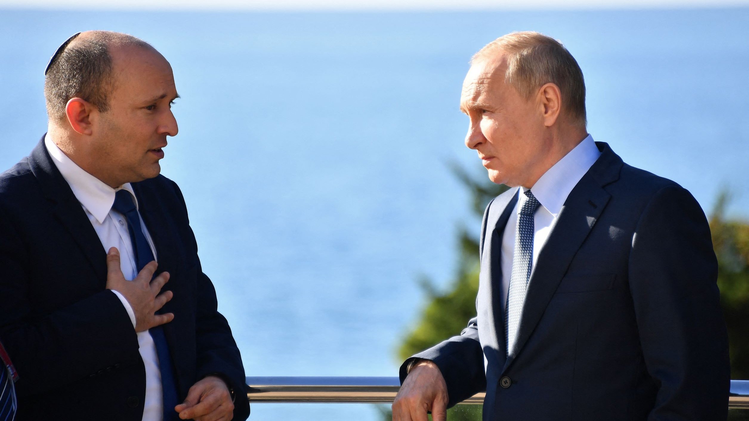 تظهر الصورة شخصين يتحدثان معًا، يبدو أن المحادثة جادة. أحدهما يرتدي بدلة رسمية مع ربطة عنق، بينما الآخر يرتدي أيضًا بدلة ولكن مع شارة دينية على رأسه. الخلفية تظهر منظرًا طبيعيًا جميلًا مع البحر. يُظهر كل منهما اهتمامًا بما يقوله الآخر، مما يوحي بأهمية النقاش بينهما.