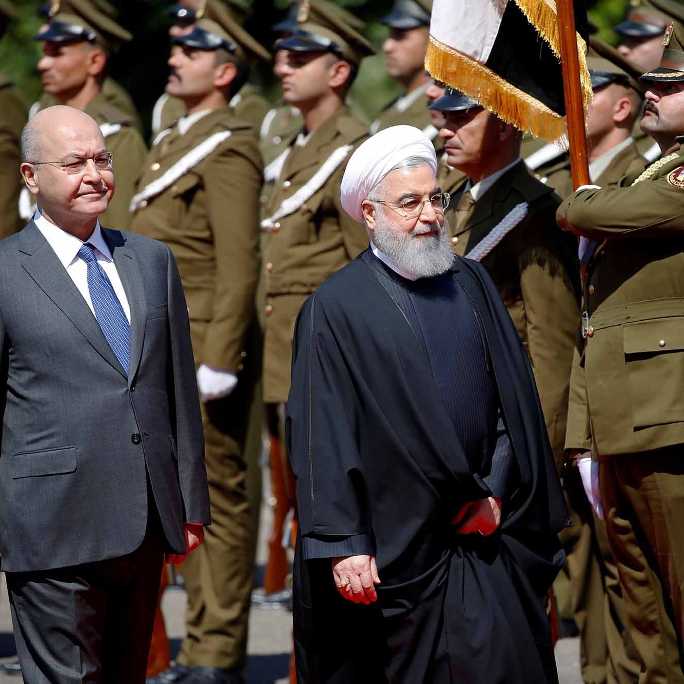 الصورة تظهر شخصيتين سياسيين يسيران معًا، حيث يبدو أنهما في مراسم رسمية. الشخص الأول يرتدي بدلة رسمية، بينما الشخص الثاني يرتدي زيًا تقليديًا أسود مع عمامة. خلفهم يظهر حرس الشرف يرتدون uniforms عسكرية، ويحملون الأعلام. الأجواء تعكس طابعًا رسميًا وجديًا، كما أن الإضاءة واضحة مما يدل على أنها تم التقاطها خلال النهار.