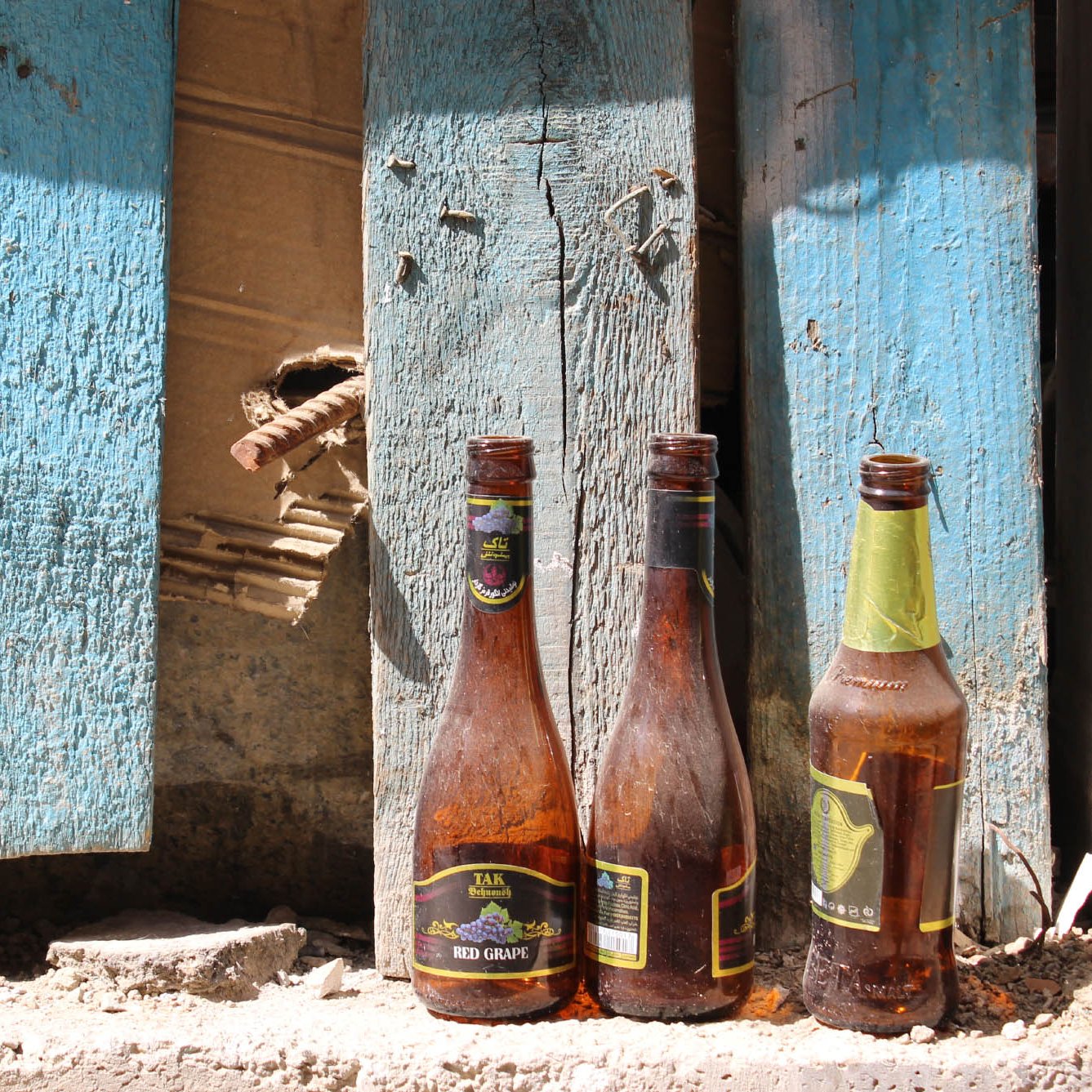 L'image montre trois bouteilles en verre vides, posées sur un sol poussiéreux. Elles sont de formes et de tailles différentes, et montrent des traces de saleté et d'usure. En arrière-plan, on aperçoit des planches de bois bleuies, dont certaines semblent endommagées, avec un trou visible. L'ensemble dégage une impression de désordre et de négligence, suggérant un espace abandonné ou en déclin.