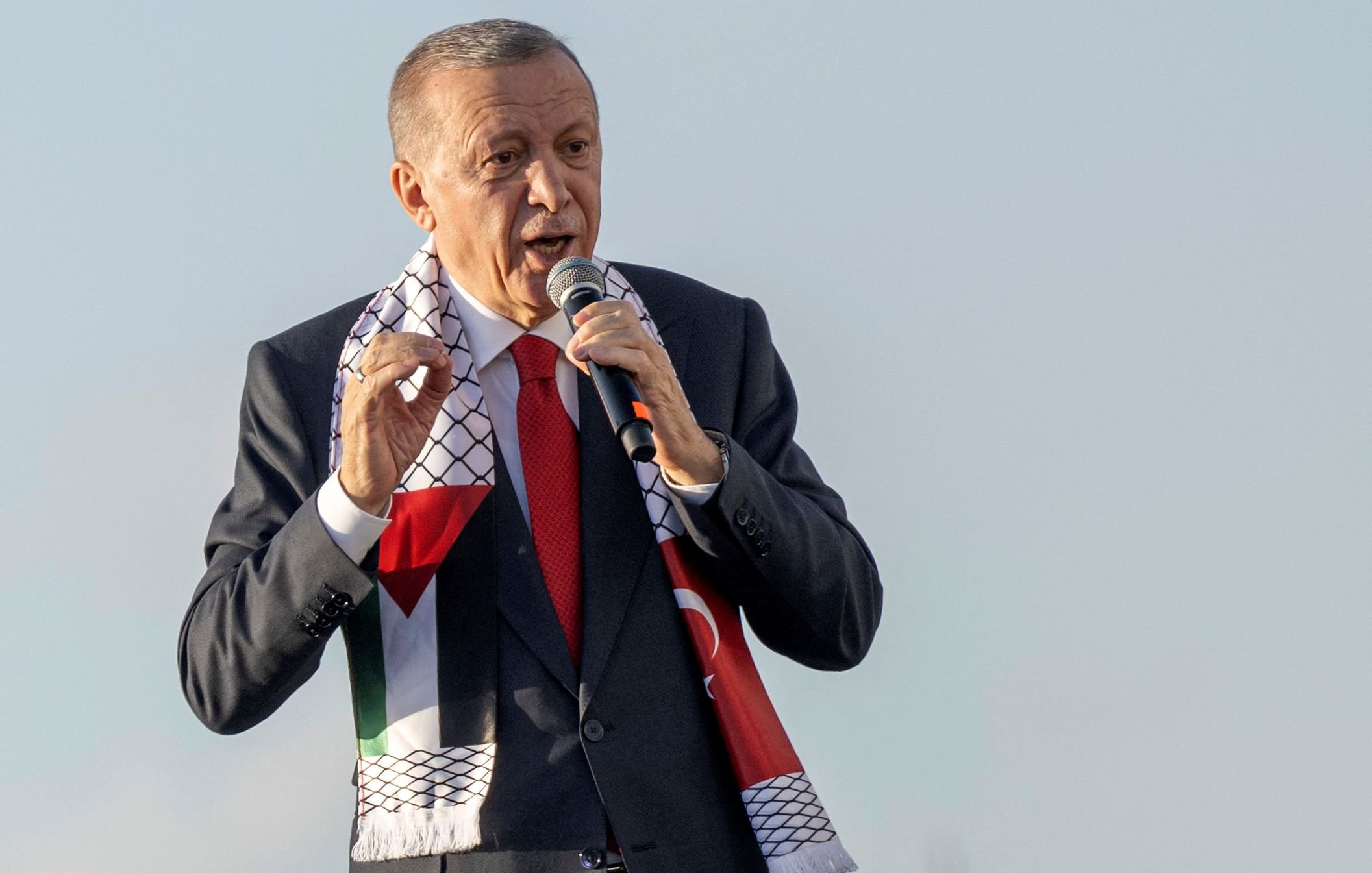 L'image montre un homme en train de parler au micro, probablement lors d'un discours ou d'une manifestation publique. Il porte un costume sombre avec une cravate rouge et un foulard aux motifs représentant la Palestine, qui est enroulé autour de son cou. L'arrière-plan semble clair, suggérant qu'il se trouve à l'extérieur. L'homme semble exprimer des émotions fortes, ce qui indique qu'il est engagé dans son discours.