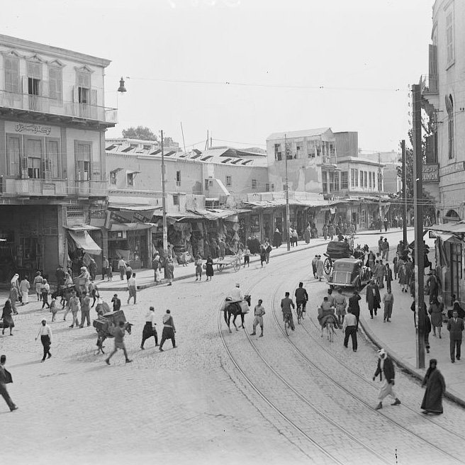 L'image montre une vue animée d'une rue commerçante dans une ville. On y voit des bâtiments à plusieurs étages avec des balcons, des étals et des commerces le long de la rue. De nombreuses personnes, y compris des piétons et des animaux, circulent. On aperçoit également une voiture ancienne sur la route. L'atmosphère semble vibrante et typique d'une époque passée, avec un mélange de textures urbaines et de la vie quotidienne.