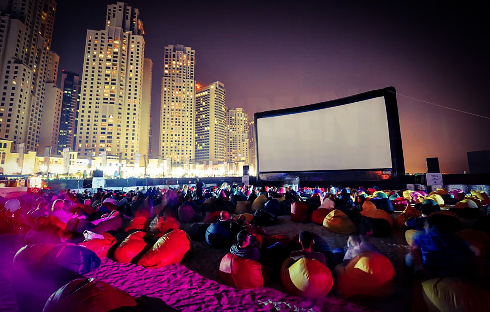 L'image montre une scène de cinéma en plein air sur une plage, animée pendant la nuit. Un grand écran est installé, et des groupes de personnes s'asseyent sur des poufs colorés, principalement de couleur rouge. En arrière-plan, on peut voir des gratte-ciels illuminés, créant une atmosphère vibrante et moderne. L'éclairage doux et les ombres ajoutent à l'ambiance festive du lieu.
