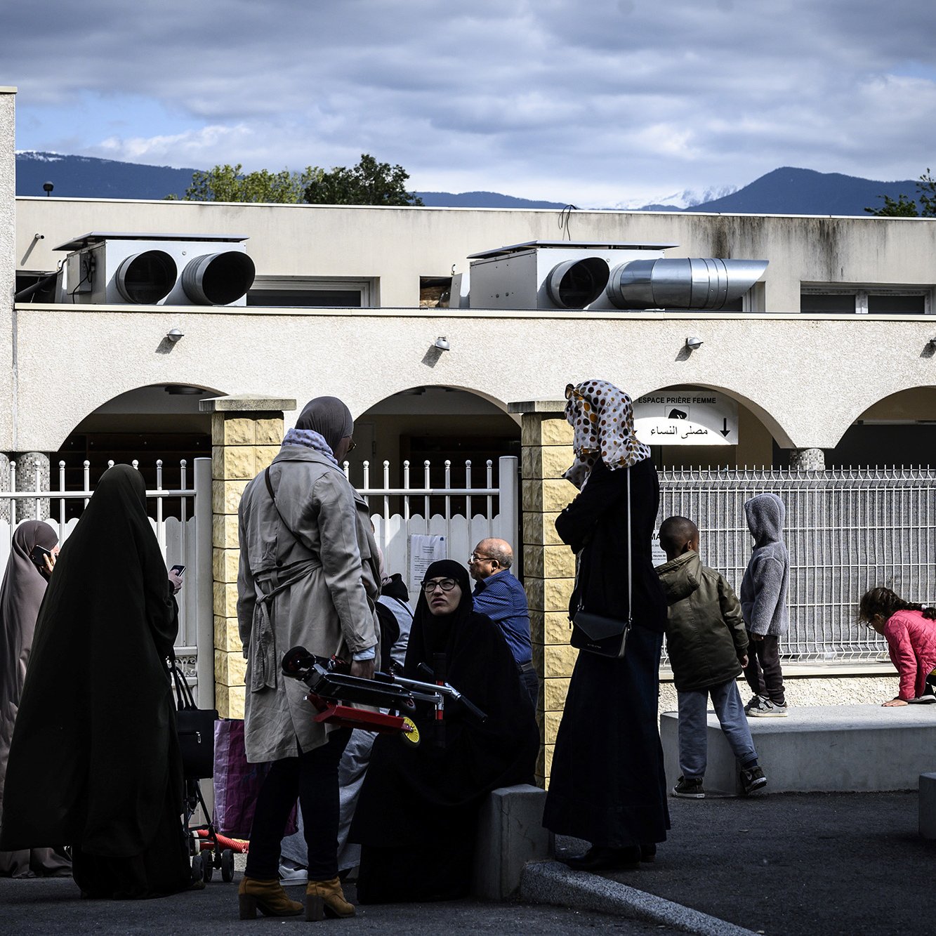 The image depicts a scene outside a building with several people present. Some individuals are wearing traditional clothing, including hijabs, while others are dressed in more casual attire. In the background, there are air conditioning units on the building, and a few trees and mountains can be seen in the distance. The setting appears to be a community space or public area, with a mix of adults and children interacting with one another. The atmosphere seems calm and social.