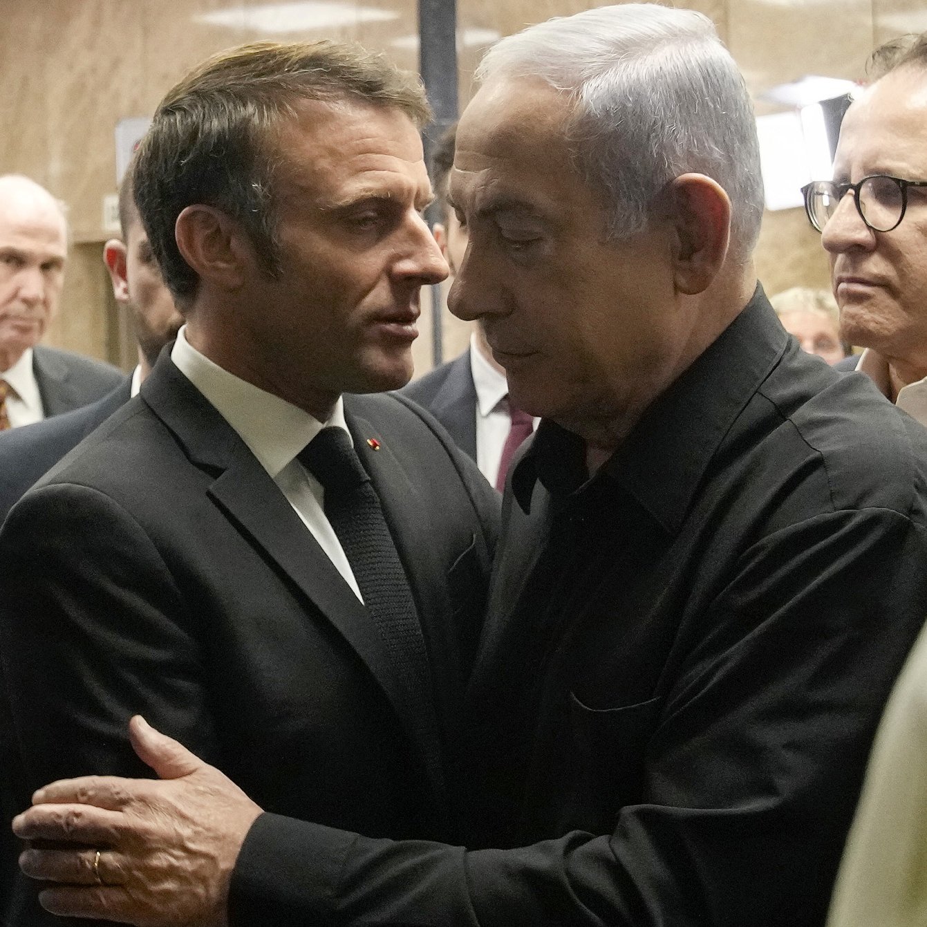 L'image montre deux hommes en discussion proche l'un de l'autre. L'un est en costume noir et le second porte une chemise noire. Ils semblent engageants dans une conversation sérieuse, entourés d'autres personnes en arrière-plan. L'ambiance générale évoque une rencontre formelle ou un événement important.
