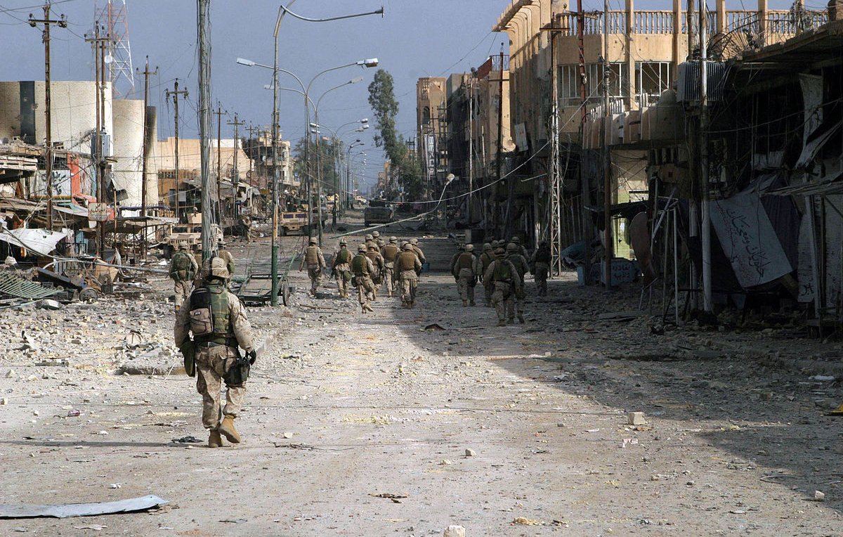 L'image montre une scène urbanisée dévastée, probablement dans une zone de conflit. On y voit des soldats marchant sur une rue déserte, entourés de bâtiments partiellement détruits. Le sol est jonché de débris, et l'atmosphère semble chargée de tension et de désolation. Les structures autour d'eux, avec des fils électriques en désordre, témoignent des ravages causés par le conflit. La lumière est diffuse, ce qui renforce le sentiment d'un environnement difficile et chaotique.