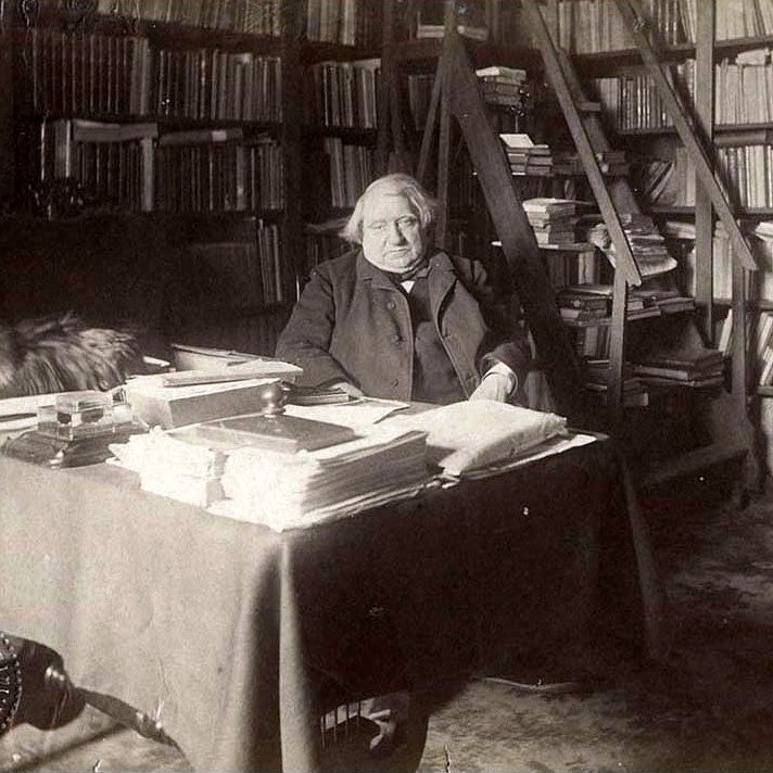 L'image montre une scène d'intérieur, probablement dans un bureau ou une bibliothèque. Un homme, apparemment âgé, est assis à une table chargée de papiers et de livres. Derrière lui, on peut voir des étagères remplies de livres. Il y a également un tapis sur le sol. Un chat, qui semble être sur la table ou à proximité, ajoute une touche de vie à l'atmosphère studieuse de la pièce. L'éclairage donne une impression de sérénité et de calme.