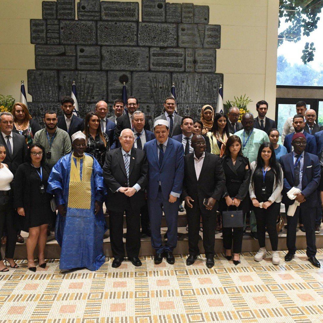 تظهر الصورة مجموعة كبيرة من الأشخاص الذين يقفون معاً في مكان مغلق. يرتدي البعض ملابس رسمية، بينما يرتدي الآخرون ملابس تقليدية. يوجد خلفهم علم أو شعار يشير إلى وهو مرتكز على جدار مزخرف. يبدو أن الحدث يجمع أفراداً من خلفيات متنوعة، ويمثل مجموعة من المشاركين في مناسبة رسمية أو مؤتمر. تعكس الصورة تفاعلًا إيجابيًا بينهم.