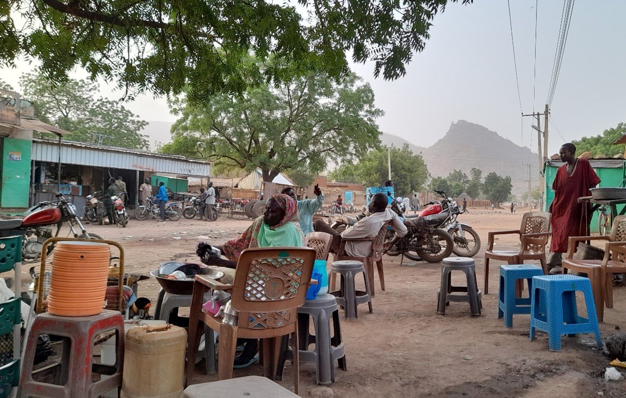 L'image montre une scène de la vie quotidienne dans un village. On y voit plusieurs chaises de différents styles et couleurs, dont certaines sont empilées. À l'arrière-plan, il y a des maisons, des collines, et plusieurs motos. La végétation est présente avec quelques arbres, offrant de l'ombre. On aperçoit également des personnes assises et se déplaçant, ce qui donne un aperçu de l'ambiance locale. Les routes sont poussiéreuses, suggérant une atmosphère rurale.
