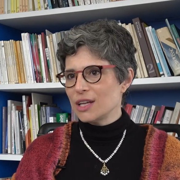 L'image montre une femme avec des cheveux courts et grisonnants, portant des lunettes et un pull noir. Elle est assise devant une bibliothèque remplie de livres, avec un mur bleu derrière elle. Elle a un collier en perles et semble être en train de parler ou de répondre à une question, affichant une expression réfléchie.