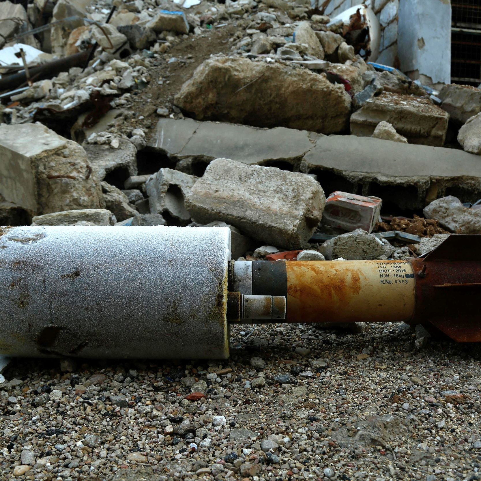L'image montre un obus ou une roquette qui est reposé sur un sol poussiéreux, entouré de débris et de morceaux de béton. En arrière-plan, on peut voir un paysage marqué par des ruines, suggérant une scène de destruction. L'objet semble endommagé et perdu au milieu de l'effondrement des structures environnantes.