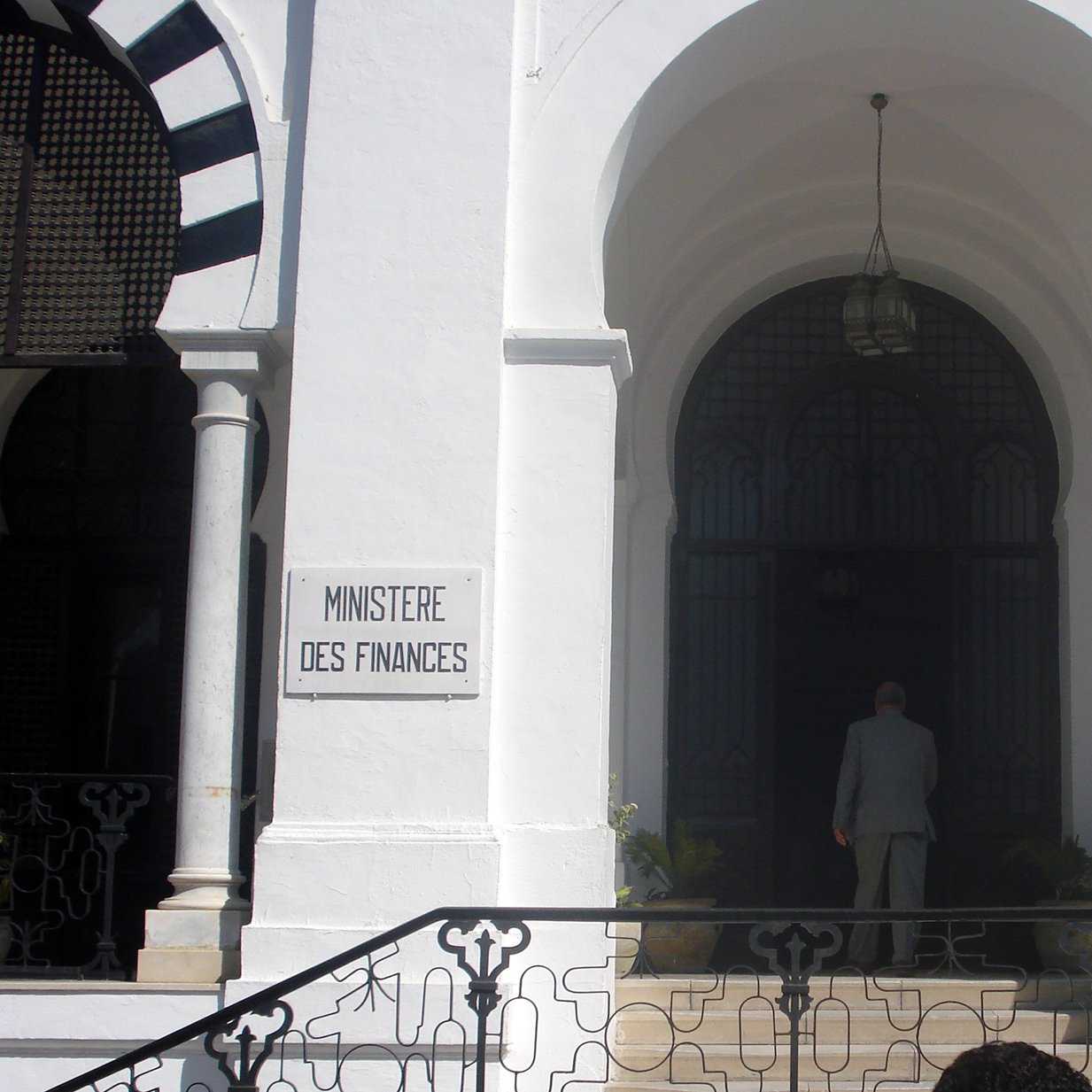 تظهر الصورة مدخل مبنى حكومي يُعرف باسم "وزارة المالية". يتميز المبنى بتصميم معماري جذاب، حيث يمكن رؤية الأبواب الكبيرة والنوافذ المزخرفة. المدخل محاط بأعمدة وأقواس، مما يعطيه طابعاً تقليدياً. هناك لافتات على الجانبين، واحدة باللغة الفرنسية والأخرى بالعربية، تشير إلى اسم الوزارة. يُلاحظ أيضاً شخص يقف أمام المدخل، مما يعكس النشاط في هذا المكان.