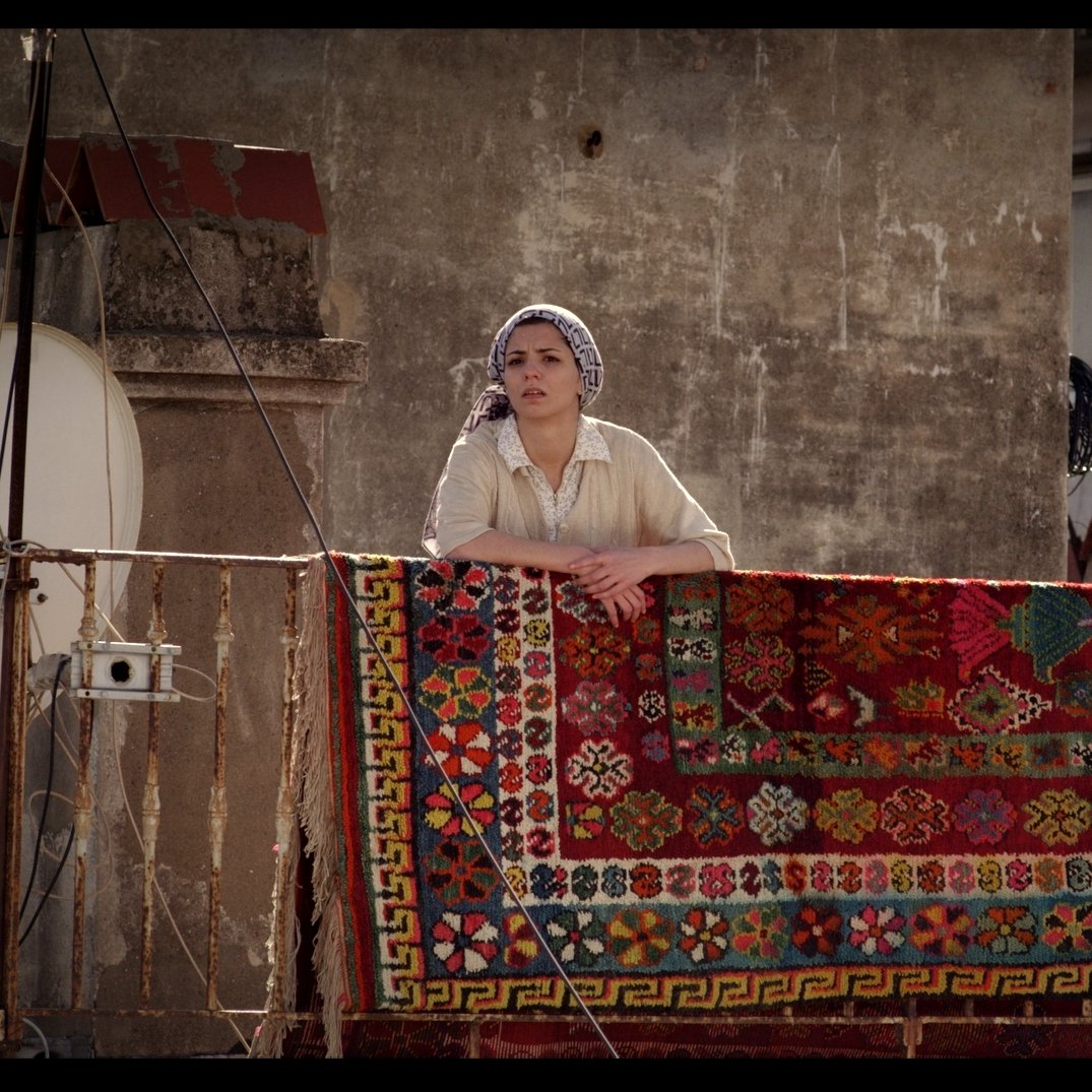 L'image montre une femme se tenant sur un balcon, regardant au loin. Elle porte un haut clair et un foulard sur la tête. Derrière elle, un grand tapis coloré est accrochée à la balustrade, avec des motifs détaillés et vibrants. En arrière-plan, on aperçoit un mur et une antenne satellite, ce qui suggère un environnement urbain. L'éclairage donne une atmosphère chaleureuse à la scène.