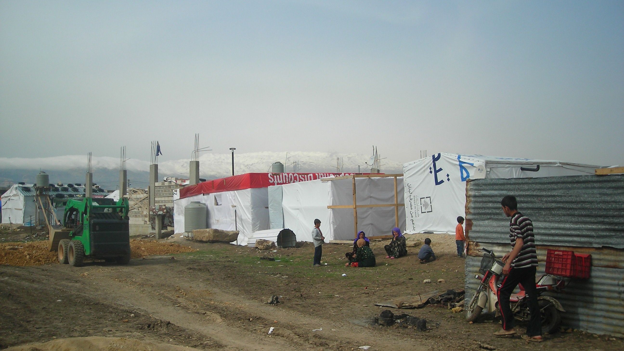 L'image montre un camp de réfugiés ou de personnes déplacées, avec des tentes blanches et des structures temporaires. Il y a un tracteur ou un véhicule de service vert à gauche. Plusieurs personnes sont visibles dans le camp, certaines assises ou debout près des tentes. Le terrain est poussiéreux, et en arrière-plan, on peut apercevoir des montagnes ou des collines. L'ensemble du paysage semble austère et reflète les conditions de vie des occupants. L'atmosphère paraît tranquille, bien que l'environnement soit rudimentaire.