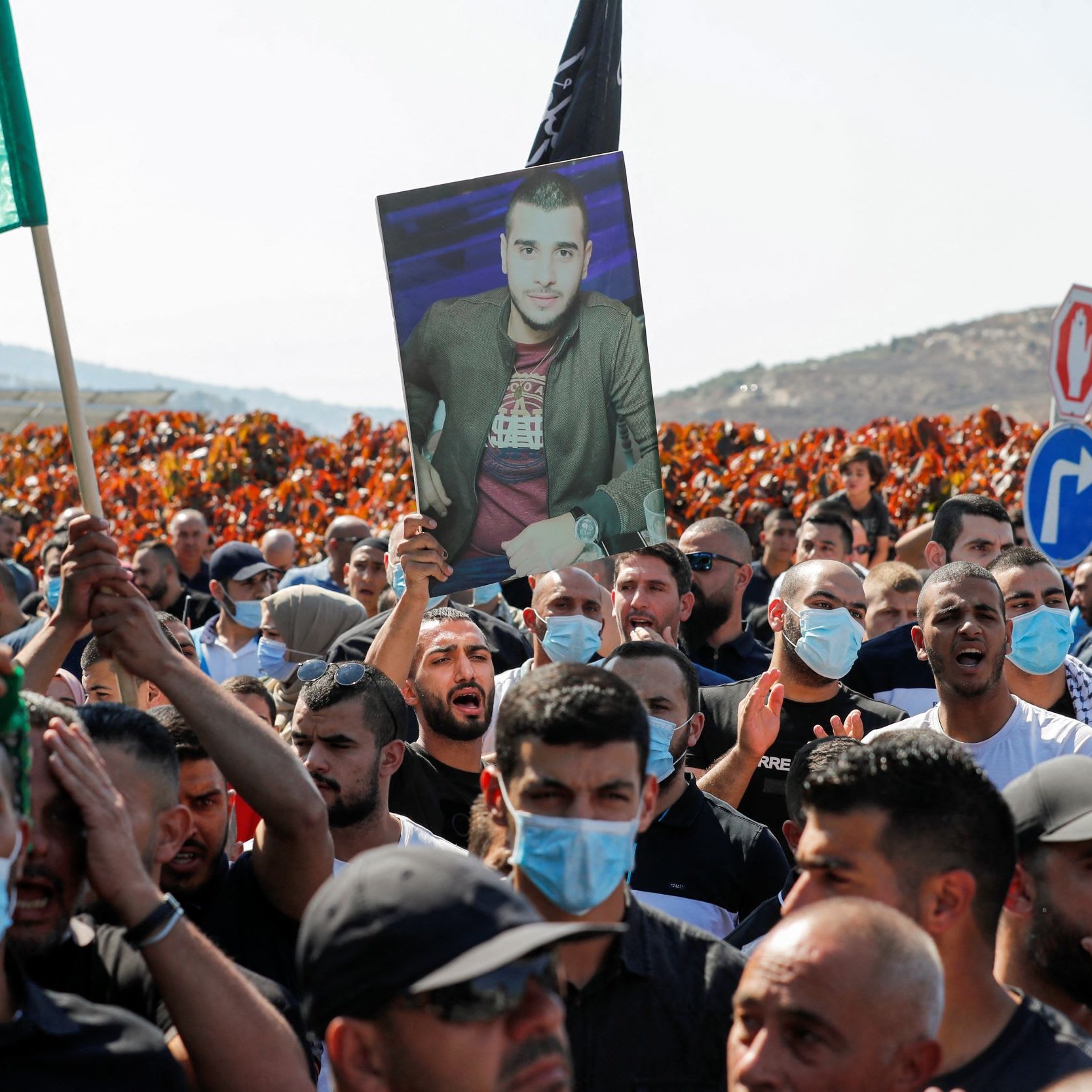 L'image montre une manifestation où une foule dense est présente. Au premier plan, des manifestants tiennent des drapeaux, dont un vert, et certains agitent des pancartes. Parmi eux, une personne porte un grand portrait d'un individu, visiblement en hommage ou en soutien. Les manifestants semblent engagés et déterminés, et ils portent tous des masques sanitaires, indiquant un contexte de pandémie. En arrière-plan, des collines se dessinent, avec des panneaux de signalisation visibles. L'atmosphère semble chargée d'émotion et de revendications.