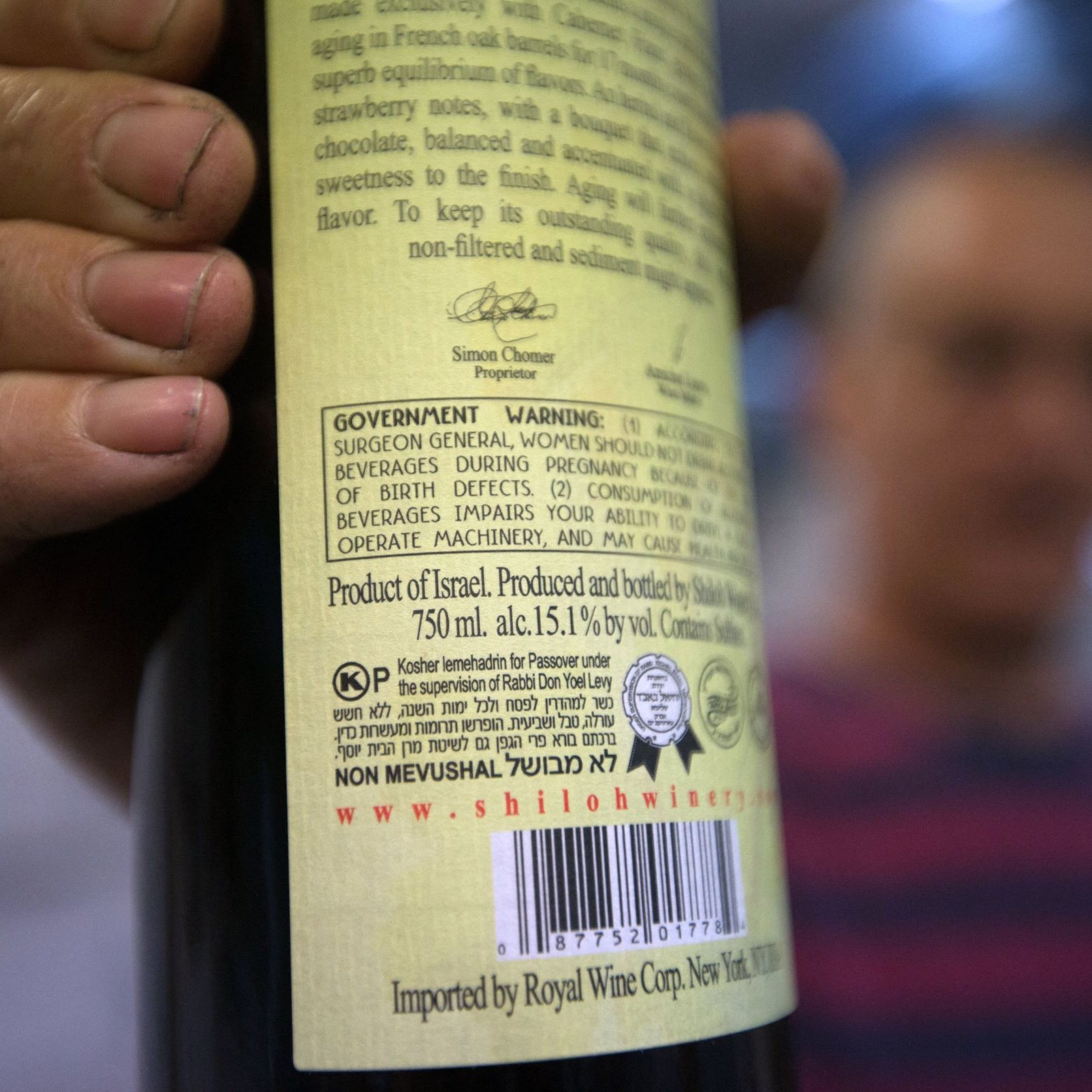 L'image montre une personne tenant une bouteille de vin, avec l'étiquette clairement visible. L'étiquette comporte des informations détaillées sur le produit, y compris des avertissements et des mentions sur la provenance, indiquant qu'il s'agit d'un vin produit en Israël. La bouteille est de 750 ml et a un taux d'alcool de 15,1 % vol. En arrière-plan, on peut deviner une autre personne floue, mais les détails ne sont pas très clairs. Les mains de la personne tenant la bouteille semblent légèrement sales, ce qui pourrait suggérer un environnement de travail ou une activité en plein air.