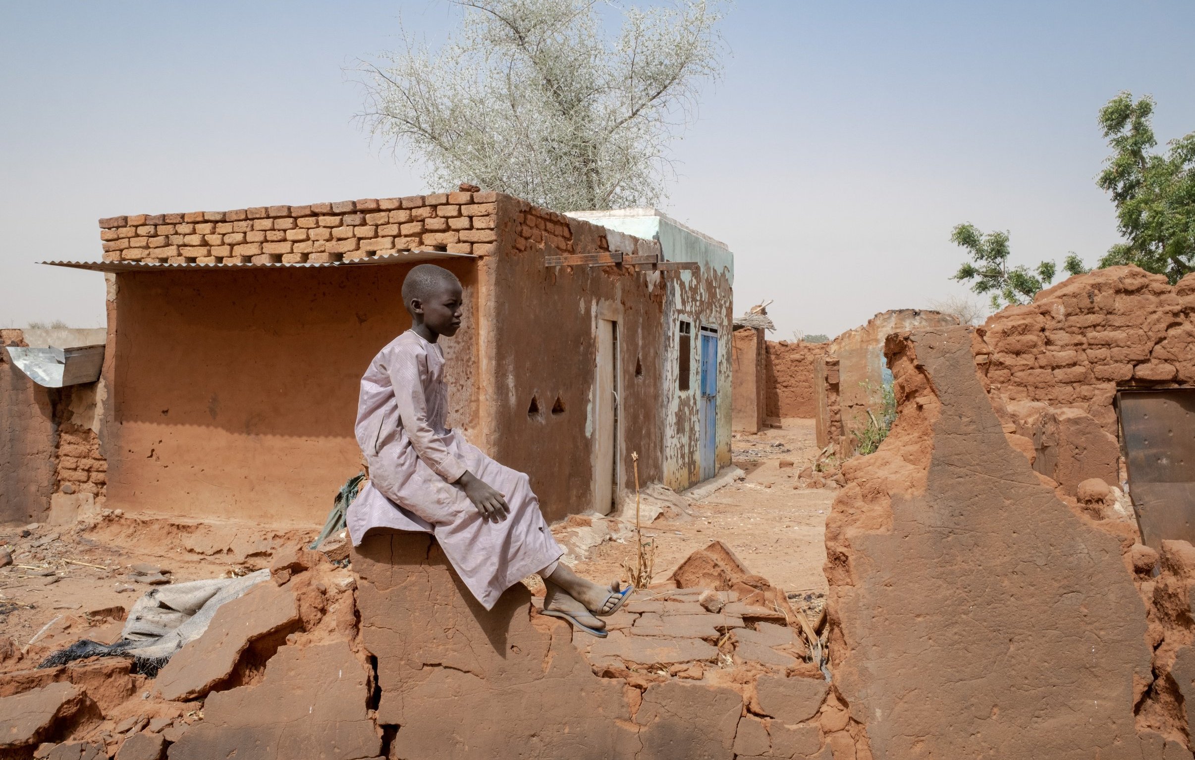 L'image montre un jeune garçon assis sur de décombres dans une région désertique. Il porte une tunique traditionnelle et semble pensif. En arrière-plan, il y a des bâtiments en terre cuite, dont certains sont partiellement détruits. Le paysage est aride, avec quelques arbres éparpillés, et le ciel est clair. L'atmosphère dégage une impression de solitude et de résilience face aux conditions difficiles.