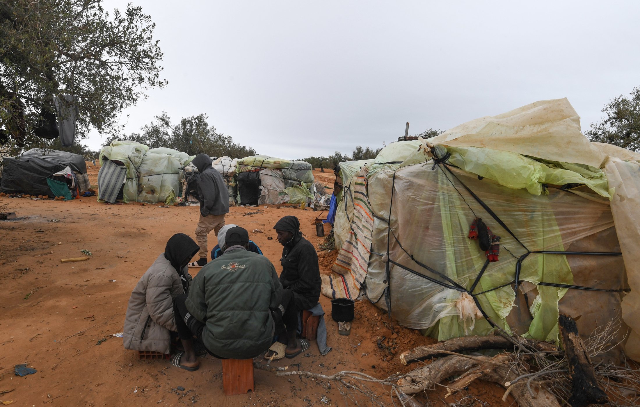 L'image montre un groupe de personnes assises ensemble sur le sol, entourées de tentes faites de matériaux recyclés, telles que des bâches en plastique. Le décor est désertique, avec un sol de sable et quelques arbres à l'arrière-plan. Les personnes semblent être en conversation ou en train de partager quelque chose. Le temps paraît nuageux, ajoutant une atmosphère sombre à la scène.