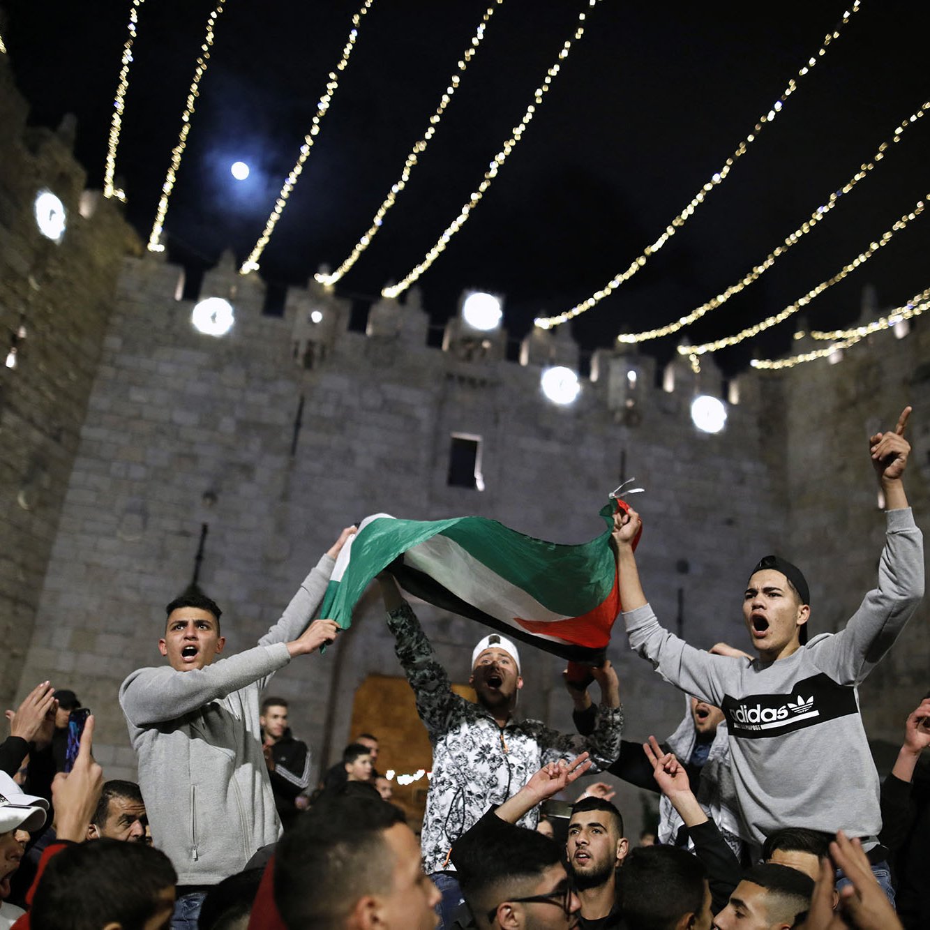 L'image montre un rassemblement nocturne devant un bâtiment ancien, probablement une fortification. Des personnes sont réunies en célébrant, certaines agitant un drapeau palestinien. L'atmosphère semble festive et dynamique, avec des lumières scintillantes qui illuminent la scène. Les expressions des participants retranscrivent une ambiance d'excitation et d'engagement.