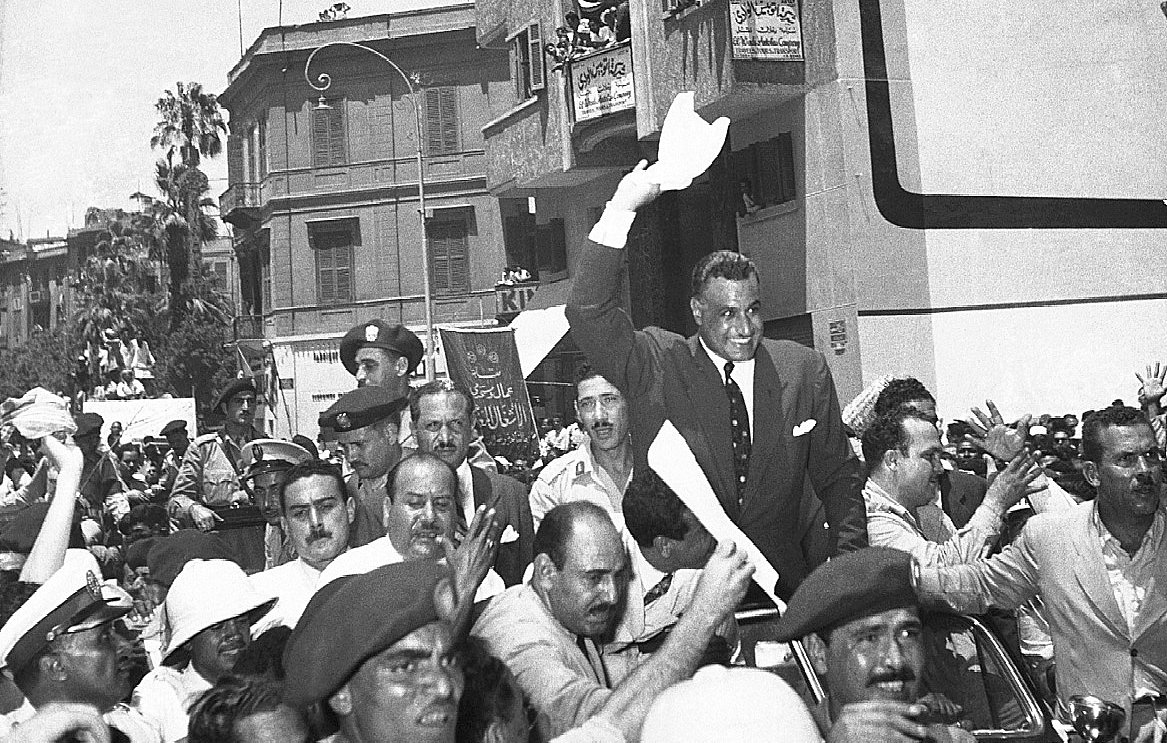 L'image montre une scène de foule animée, probablement lors d'un événement historique. De nombreuses personnes sont rassemblées autour d'une voiture, où un homme, visiblement en position d'autorité, lève les bras en signe de salutation ou d'approbation. Les spectateurs semblent enthousiastes, agitant des banderoles ou des papiers. On peut voir des policiers et des militaires en uniforme, ajoutant à l'ambiance de la scène. L'architecture des bâtiments en arrière-plan suggère une ville dynamique, probablement dans un contexte politique ou social significatif.