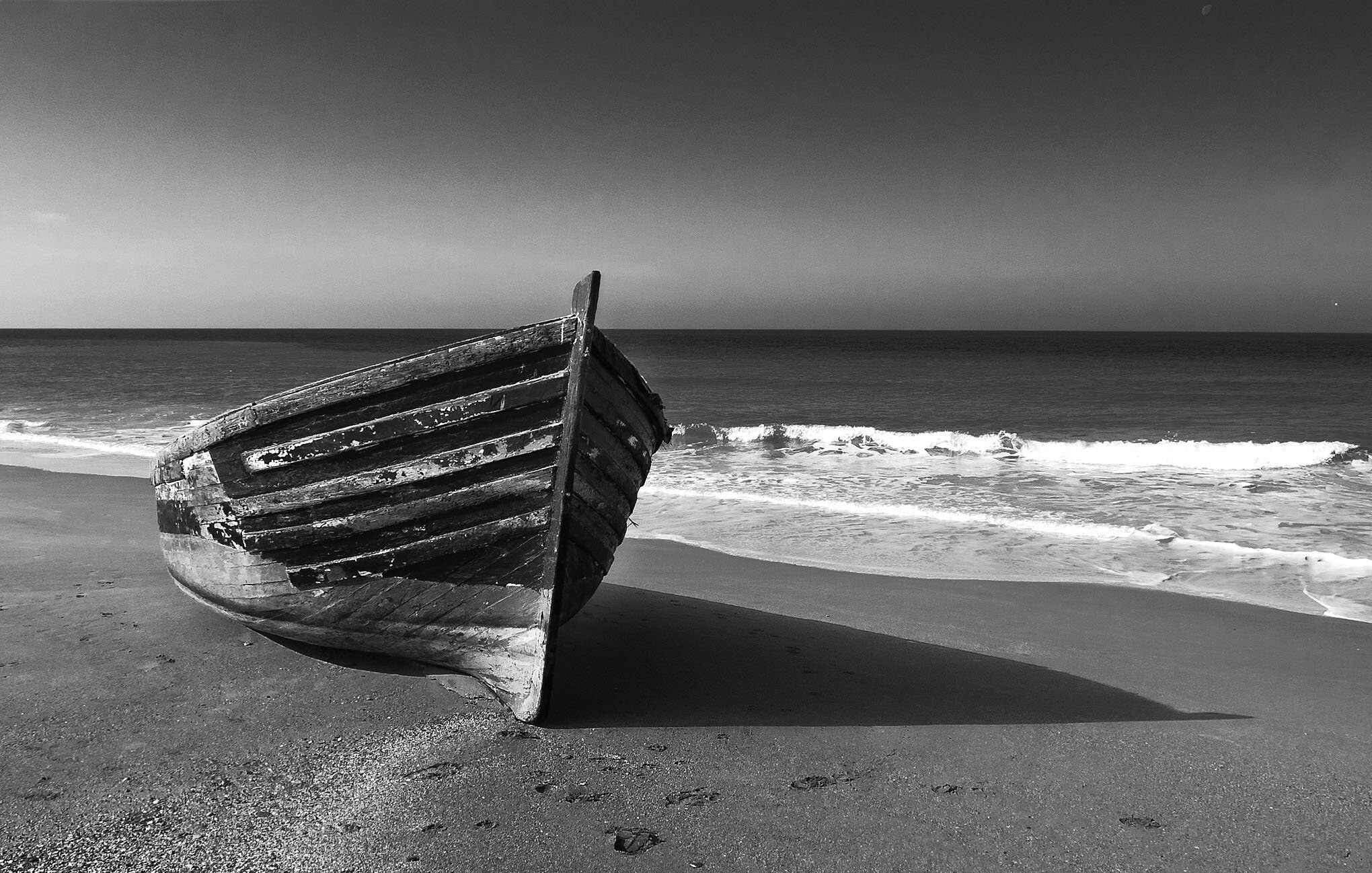 L'image montre un bateau en bois qui est échoué sur une plage. Le bateau, usé par le temps, est orienté vers la mer. La plage est accompagnée de vagues qui se brisent doucement sur le rivage. Le contraste en noir et blanc donne une atmosphère calme et mélancolique à la scène, mettant en valeur les textures du bois et les formes des vagues. Le ciel est dégagé, ajoutant à la sérénité de l'image.