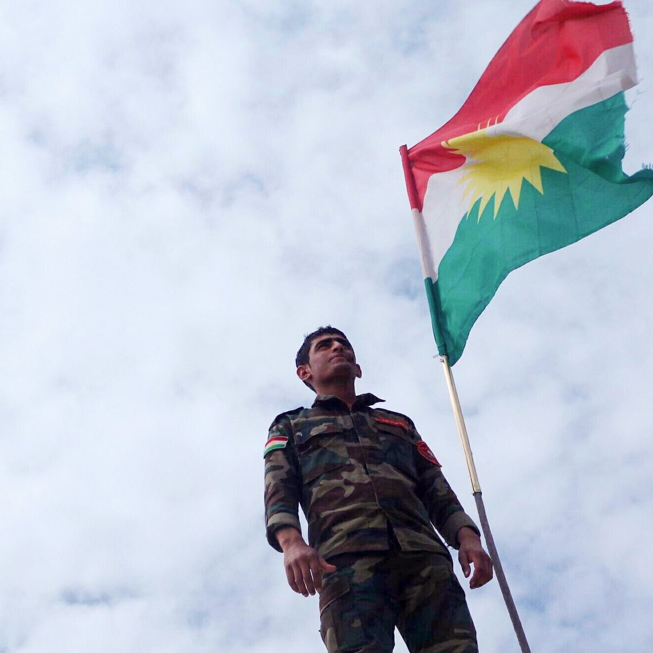 L'image montre un soldat portant un uniforme militaire, se tenant debout sous un drapeau kurde. Le drapeau présente les couleurs rouge, vert et blanc avec un soleil rayonnant au centre. Le ciel est nuageux, ajoutant une ambiance dramatique à la scène. Le soldat semble fier et déterminé, symbolisant l'engagement et l'identité kurde.