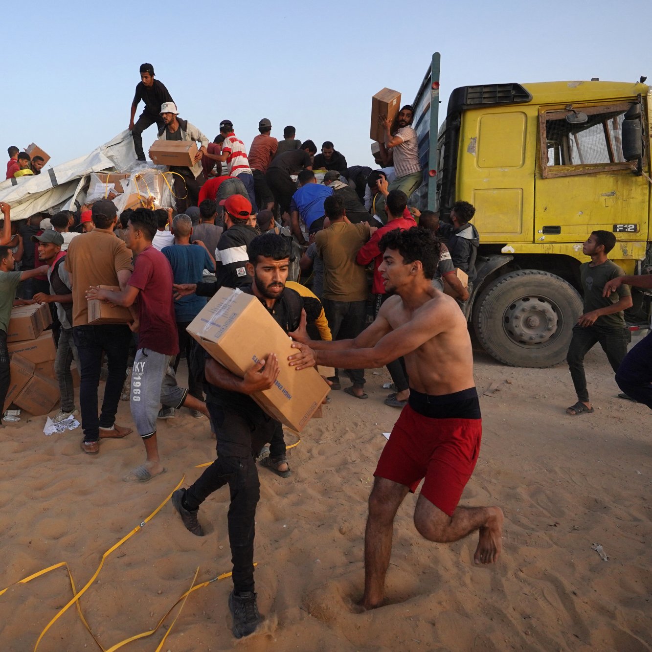 L'image montre une scène animée où de nombreuses personnes semblent s'affairer autour d'un camion jaune. Des individus portent des boîtes et se déplacent rapidement, certains courant, tandis que d'autres semblent aider à décharger le camion. L'environnement est désertique, avec du sable au sol. L'atmosphère est chargée d'activité et d'urgence, indiquant qu'ils sont probablement en train de distribuer de l'aide ou des provisions.