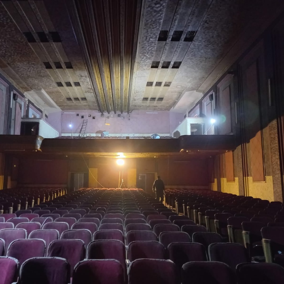 L'immagine mostra l'interno di un teatro abbandonato. Si possono vedere file di poltrone di velluto viola, di fronte a un palcoscenico che sembra vuoto. La luce proveniente da un proiettore evidenzia il pavimento e crea un'atmosfera suggestiva. Sul palcoscenico, ci sono alcune attrezzature, come scale, suggerendo che ci siano lavori in corso o recenti ispezioni. Le pareti del teatro mostrano segni di degrado, contribuendo a un senso di malinconia e nostalgia.