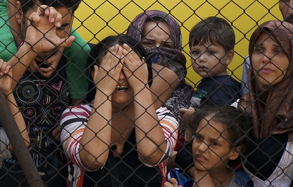 L'image montre un groupe de personnes derrière une clôture en métal. Au centre, une femme semble très émue et cache son visage avec ses mains, tandis que plusieurs enfants et adultes l'observent avec des expressions d'inquiétude et de tristesse. Le contraste entre la clôture et les visages des personnes souligne un sentiment d'angoisse et de vulnérabilité. Les couleurs de l'arrière-plan sont vives, mais l'atmosphère ressentie est lourde et chargée d'émotions.