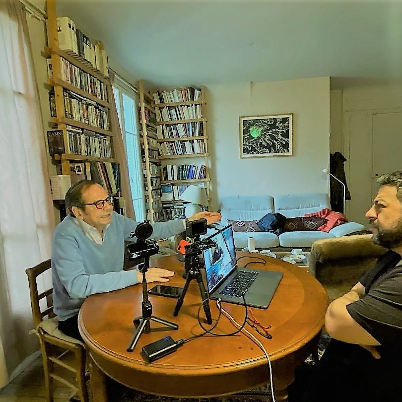 تظهر في الصورة مشهداً من داخل غرفة معيشة. على الطاولة الدائرية توجد أجهزة كمبيوتر وكاميرات، حيث يجلس شخصان يتحدثان معًا. الشخص الأول يرتدي سترة زرقاء ويبدو أنه يتحدث بفعالية، بينما الشخص الثاني يجلس مقابله، عابسًا أو متفكرًا. في الخلفية، توجد رفوف مليئة بالكتب، وأريكة مريحة، وظهر شخص آخر مستلقيًا على الأريكة. الإضاءة في المكان تبدو طبيعية، مما يعطي شعورًا بالراحة والتركيز على الحوار.