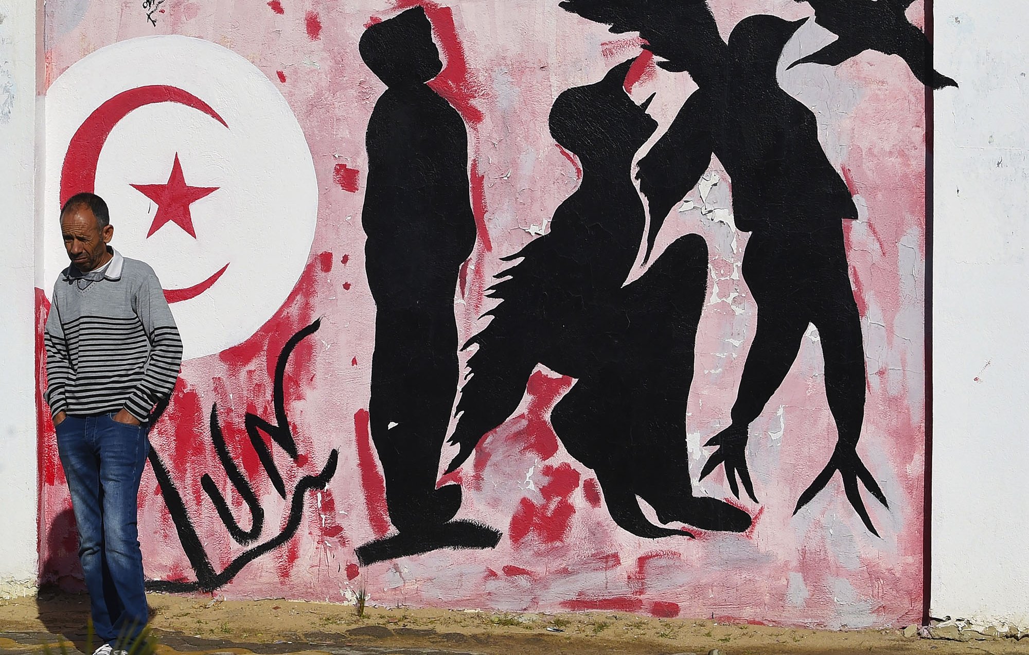 L'immagine mostra un murale su un muro bianco, caratterizzato da una palette di colori rossi e neri. Sullo sfondo, è presente un grande simbolo della luna crescente e una stella, simboli spesso associati alla bandiera tunisina. Al centro, ci sono delle silhouette di figure umane che appaiono in diverse posizioni: alcune sono in piedi, altre sembrano inginocchiarsi o muoversi in modo dinamico. In primo piano, un uomo con una maglietta a strisce passeggia accanto al murale, creando un contrasto tra l'arte e la vita quotidiana. L'atmosfera trasmette un senso di cultura e identità, evidentemente legato alla Tunisia.