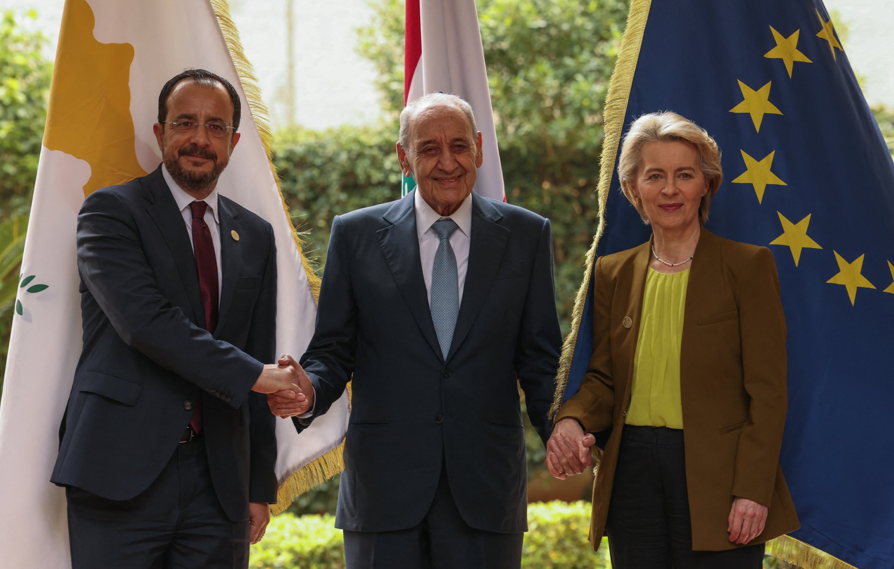 L'image montre trois personnes posant ensemble pour une photo. Au centre, un homme âgé souriant est entouré de deux personnes : à gauche, un homme en costume avec une cravate, et à droite, une femme en tenue professionnelle. Ils se tiennent la main en signe de coopération. En arrière-plan, on peut voir des drapeaux, dont un de l'Union européenne, ainsi que d'autres drapeaux nationaux. L'environnement est verdoyant et semble être en extérieur.
