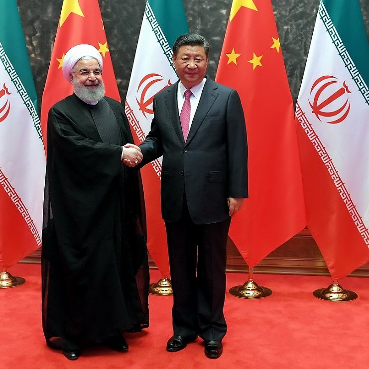 تظهر الصورة اثنين من القادة وهما يتصافحان في مكان رسمي. خلفهما توجد مجموعة من الأعلام التي تمثل كل من إيران والصين. الأعلام مرتبة بشكل يعكس التعاون بين البلدين. المشهد يبدو ودياً، حيث يرتدي أحد القادة زيّاً تقليدياً إيرانياً بينما الآخر يرتدي بدلة رسمية. الأجواء تميل إلى الاحتفالية مع سجادة حمراء وألوان زاهية للأعلام.