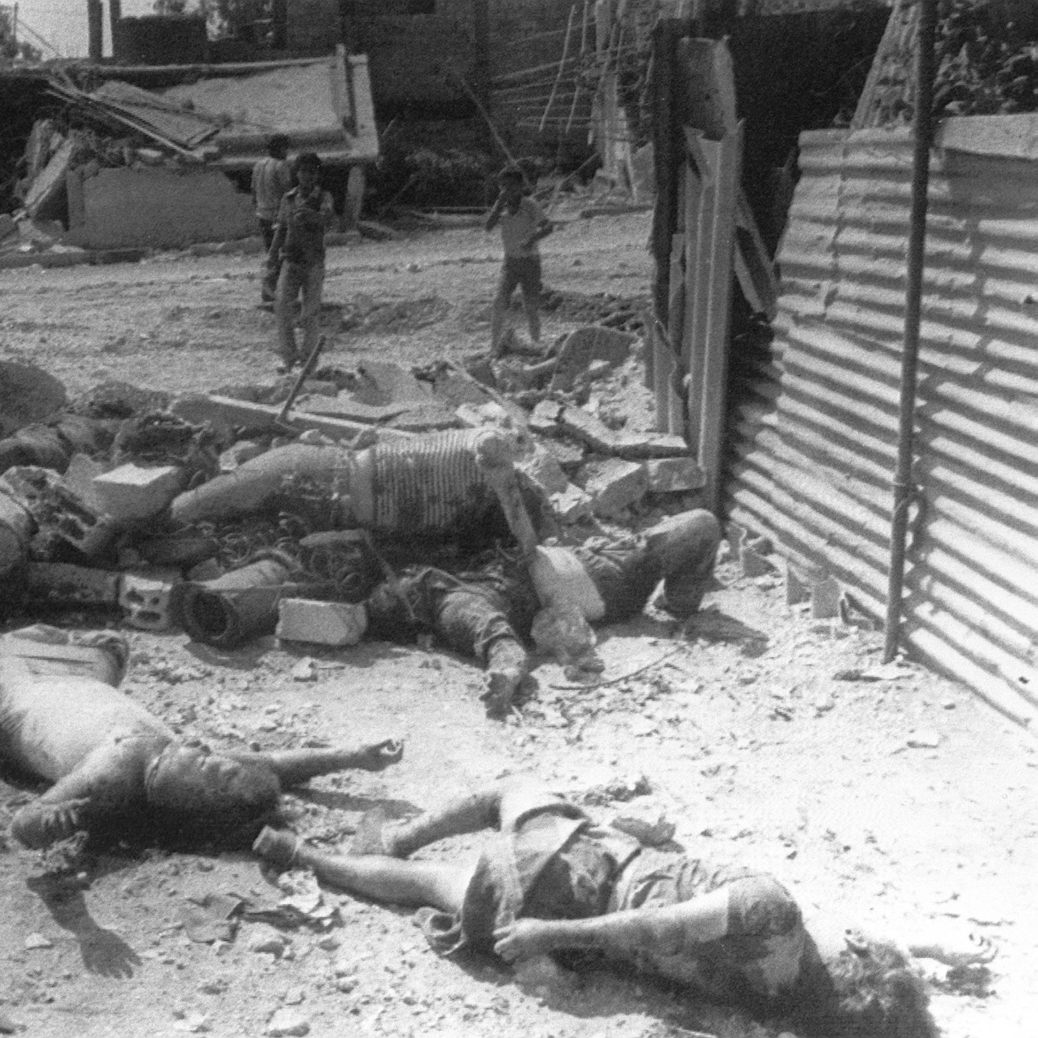 L'immagine mostra un ambiente devastato, probabilmente dopo un attacco o un evento distruttivo. Sul terreno ci sono corpi senza vita, apparentemente tra le macerie di edifici distrutti. In lontananza, alcune figure umane sembrano muoversi tra i detriti. La scena evoca un forte senso di sofferenza e desolazione, riflettendo gli effetti devastanti della violenza o di un conflitto.