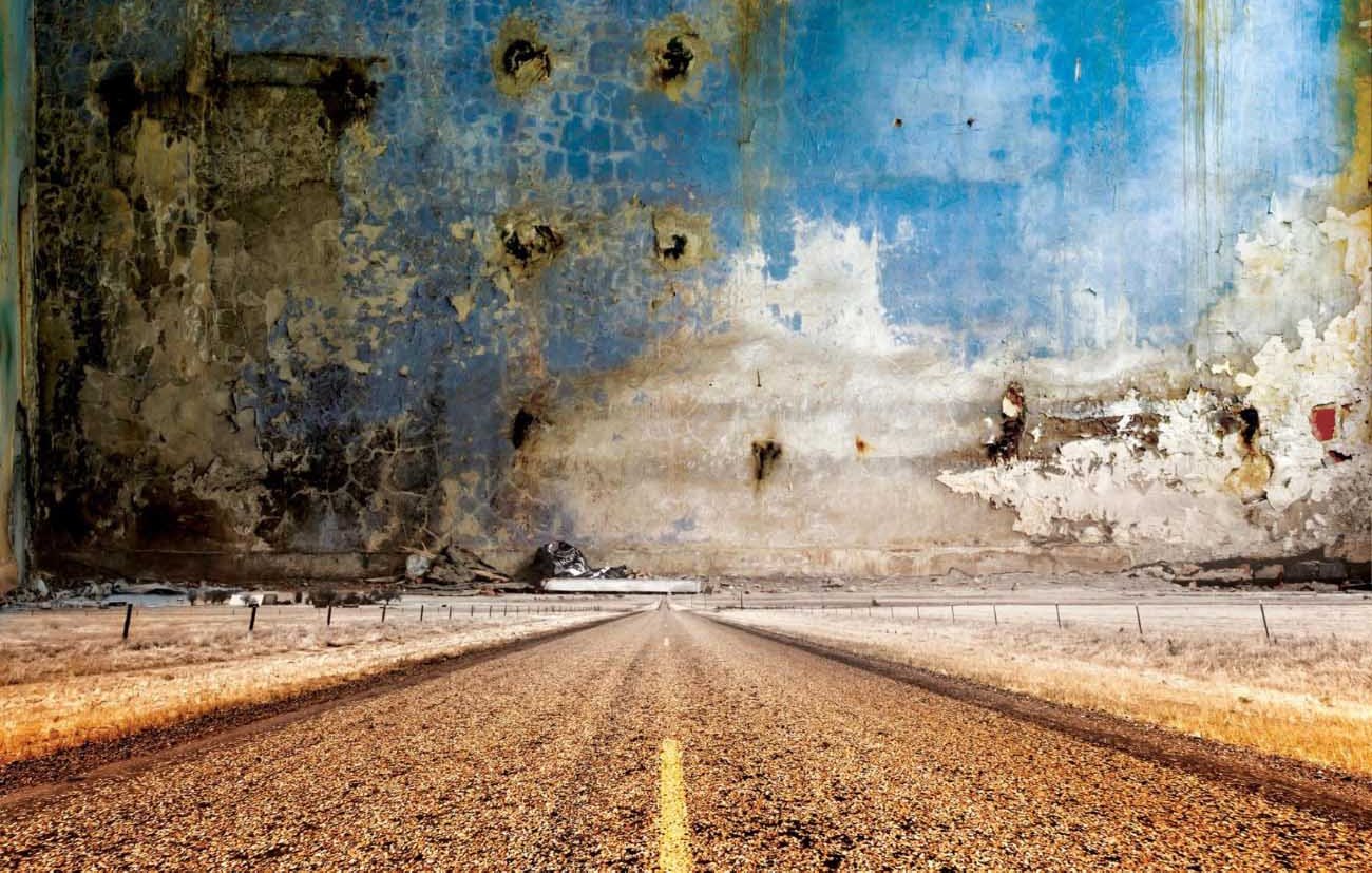 L'image montre une route qui s'étend à l'horizon, traversant un paysage désertique. De chaque côté de la route, il y a des champs secs aux teintes jaunâtres. En arrière-plan, un grand mur au look usé et décoloré s'élève, présentant des traces de peinture écaillée et des taches, probablement en raison de l'humidité ou de l'érosion. Le ciel au-dessus est d'un bleu vif, contrastant avec les teintes terreuses du sol et du mur. L'ensemble de la scène évoque une atmosphère de solitude et d'abandon.