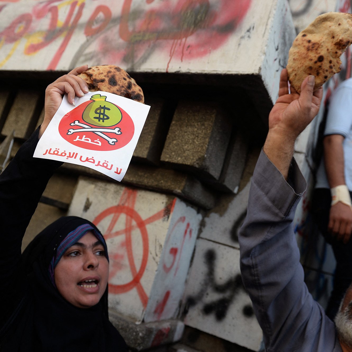 The image depicts a protest scene where individuals are holding up flatbreads and a sign. The sign features a dollar symbol with a prohibition sign over it, conveying a message against inflation or economic hardship, likely related to rising food prices. In the background, there are graffiti-covered walls, indicating a tense atmosphere. The protesters express their discontent, while another person appears in the background observing the scene.