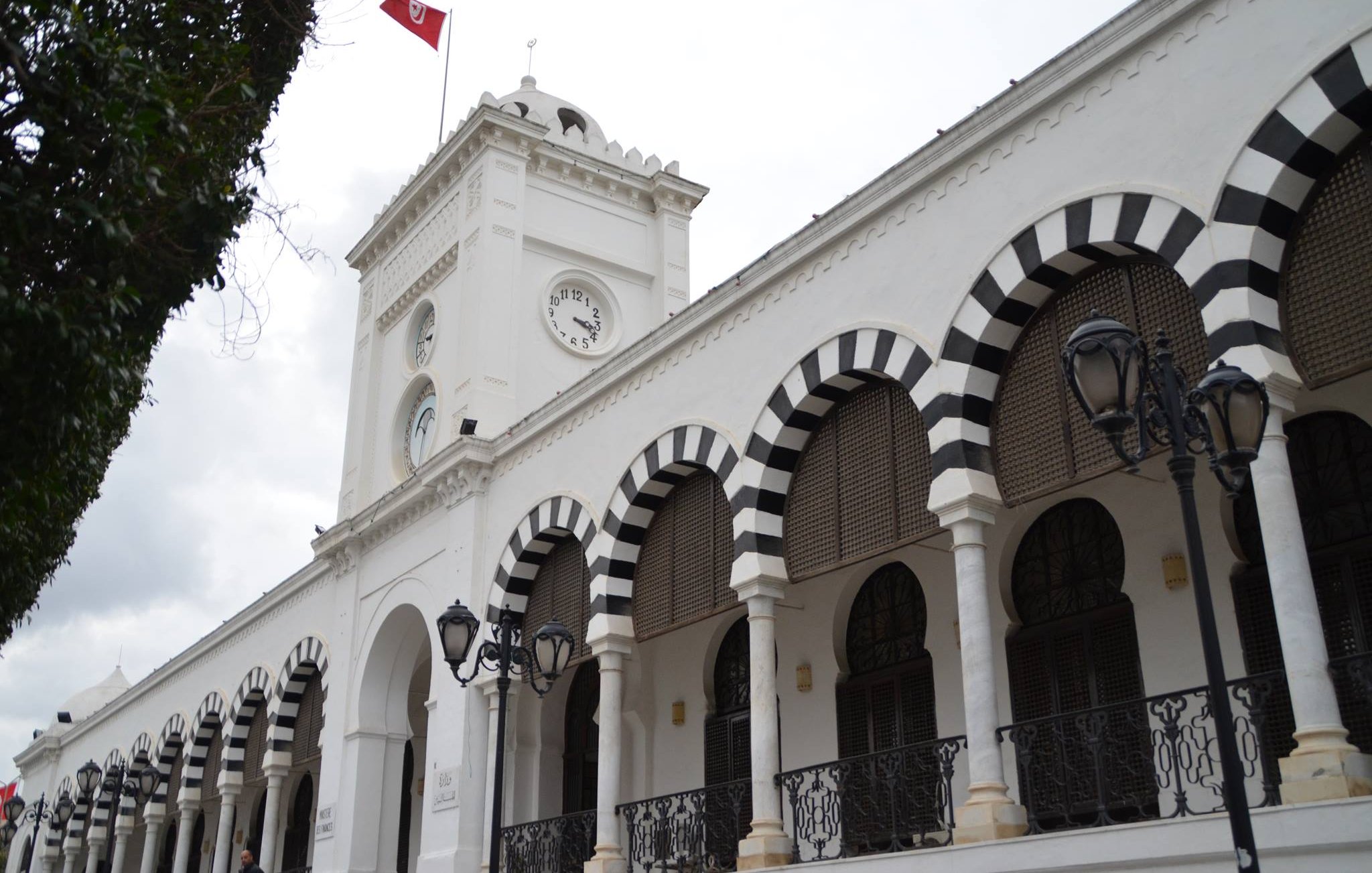 L'image montre un bâtiment architectural blanc avec des arcades noires et blanches. Il y a une horloge sur la façade et un drapeau tunisien flottant au sommet. Le ciel est nuageux, ce qui donne une ambiance légèrement sombre. Des lampadaires sont visibles le long de la structure, ajoutant une touche d'élégance à l'ensemble.