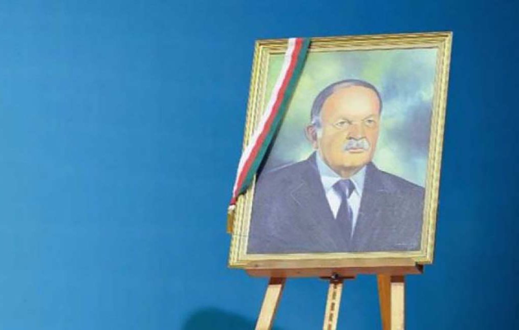 L'image montre un portrait encadré d'un homme, affiché sur un chevalet. Le fond est d'un bleu uniforme, créant un contraste avec le cadre doré du portrait. L'homme dans l'image a une expression sérieuse et porte un costume, accompagné d'un insigne ou d'un drapeau sur le cadre, possiblement représentant un symbole national ou officiel.