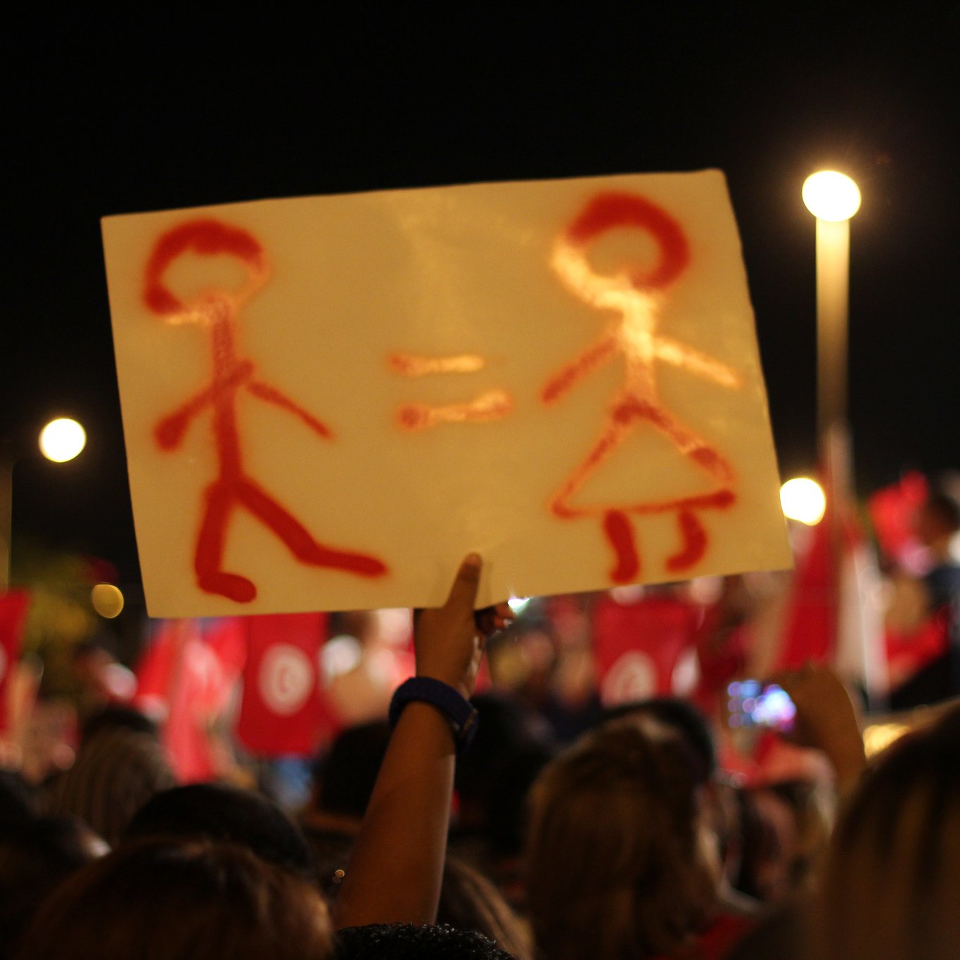 The image shows a night scene of a demonstration or rally, where a person is holding up a sign. The sign features a simplistic representation of a male figure and a female figure, with an equals sign between them, suggesting a message of gender equality. In the background, a crowd can be seen, along with red flags that likely represent a political or social movement. The atmosphere appears to be vibrant and engaged, reflecting a collective message or cause.