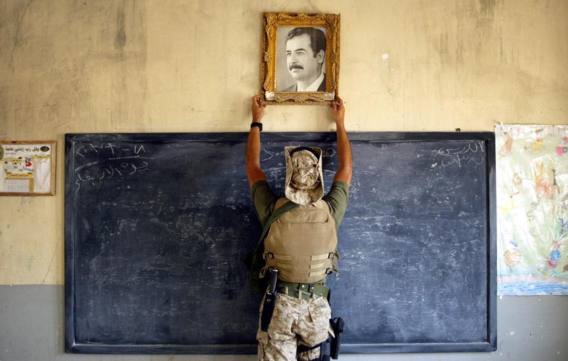 L'image montre un soldat en uniforme, portant un gilet de protection, en train de fixer un portrait dans un cadre doré sur un mur. Le portrait représente un homme aux traits marqués, probablement un dirigeant. En arrière-plan, on peut voir un tableau noir avec des inscriptions en arabe, ainsi qu'une feuille de papier accrochée au mur, qui semble être un dessin. L'atmosphère de la scène semble refléter un environnement qui a été, ou est, lié à des événements militaires ou politiques.