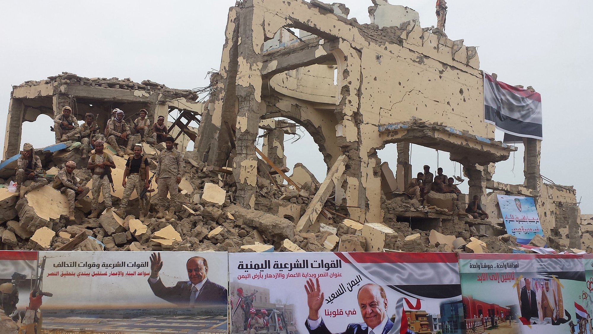 The image depicts a heavily damaged building that has partially collapsed, suggesting that it may be the result of conflict or disaster. In the foreground, there are several large banners featuring messages and images, including one that appears to be a political endorsement. Soldiers are visible on top of the rubble, indicating a military presence in the area. The overall scene conveys a sense of destruction and the impact of ongoing turmoil, likely in a war-affected region.