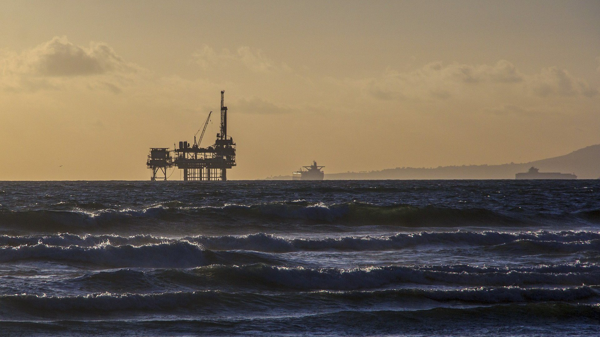 The image shows a distant oil rig standing tall in the ocean during a sunset. The sky is illuminated with soft golden hues, creating a serene atmosphere. Waves roll gently in the foreground, and in the background, a cargo ship can be seen faintly on the horizon. The overall mood is calm and peaceful, with a blend of industrial and natural elements.